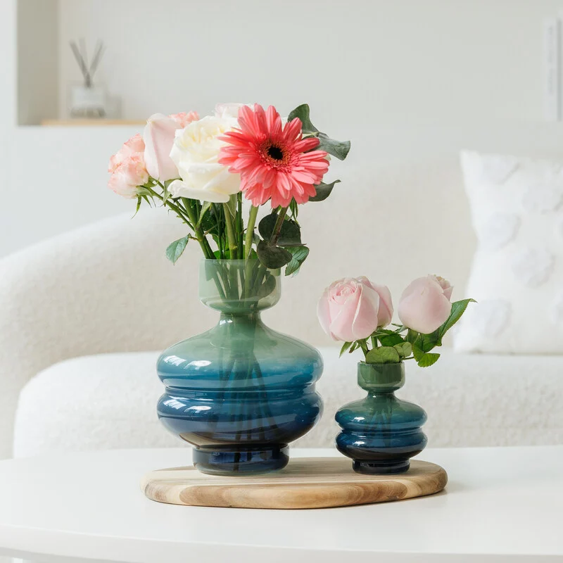 Two green-to-blue gradient Vert Nook Oddessy glass vases on a round wooden tray atop a white coffee table; the larger vase holds a bouquet of pastel roses, carnations, and eucalyptus sprigs, set against a cream sofa and minimalist white shelf in soft focus behind.